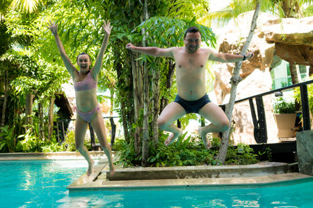 A young married couple absurdly jumping at the same time in the pool near the palm trees and laughing.の写真素材