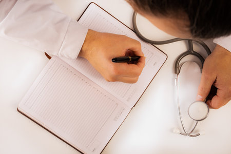 The doctor sits in an office at a white table, makes notes in a notebook, a stethoscope lies nearby, view from above. Close upの写真素材