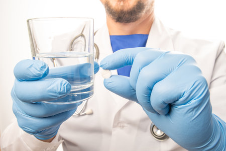 The Smile doctor in white clothes, and blue gloves, with a stethoscope on his neck, holds out pills and glass of water. Close upの写真素材