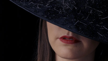 Close-up of a half-sided face of an attractive mysterious woman in a black witch hat, with bright red lips smiling. Halloween concept.の写真素材