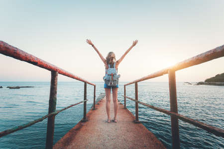 Summer landscape with young woman standing on the old sea pier with raised up arms  at sunset. Vintage toning. Girl on the seashore, turquoise water and sunny sky in the evening. Lifestyleの写真素材