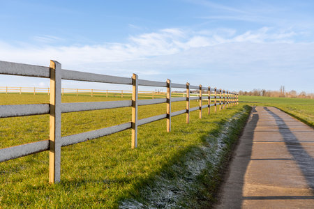 At the edge of the meadow there is wooden fence next to a road that goes down a lane through the landscapeの写真素材