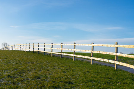 At the edge of the meadow there is wooden fence next to a road that goes down a lane through the landscapeの写真素材