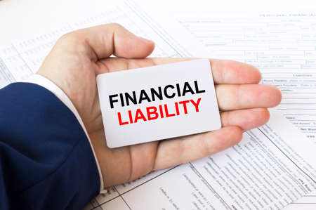 A man's hand is on the financial tables holding a business card with the inscription Financial Liability. Business concept photoの写真素材