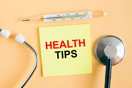 Yellow sticker with the text Health Tips. A thermometer with a phonendoscope on a warm background. Can be used as a medical concept photoの写真素材