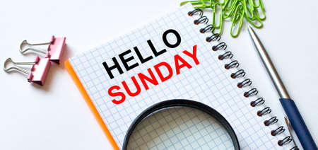 Notepad with text Hello Sunday on white background with clips, pen and magnifying glass. Can be use as concept photoの写真素材