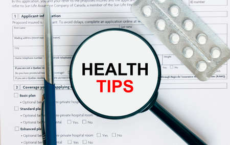 Magnifying glass with text Health Tips inside lies on medical documents with pills and a blue metal pen. Can be use as a concept photoの写真素材