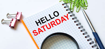 Notepad with text Hello Saturday on white background with clips, pen and magnifying glass. Can be use as concept photoの写真素材