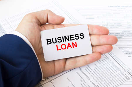A man's hand is on the financial tables holding a business card with the inscription Business Loan. Business concept photoの写真素材