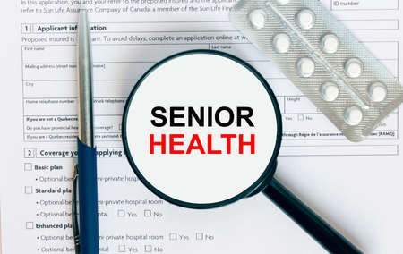 Magnifying glass with text Senior Health inside lies on medical documents with pills and a blue metal pen. Can be use as a concept photoの写真素材