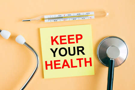 Yellow sticker with the text Keep Your Health. A thermometer with a phonendoscope on a warm background. Can be used as a medical concept photoの写真素材