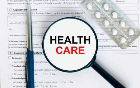 Magnifying glass with text Health Care inside lies on medical documents with pills and a blue metal pen. Can be use as a concept photoの写真素材