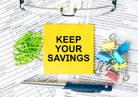 Orange sticker with text Keep Your Savings. Next to it is a blue pen with colored stationery clips, green paper clips and eyeglasse. It can be used as a business and financial conceptの写真素材