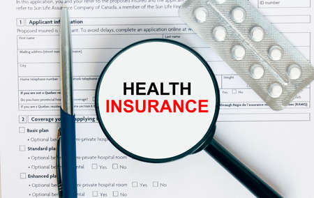 Magnifying glass with text Health Insurance inside lies on medical documents with pills and a blue metal pen. Can be use as a concept photoの写真素材