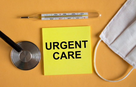 Sticker with text Urgent Care lying on the orange background with mask and thermometer. Medical conceptの写真素材