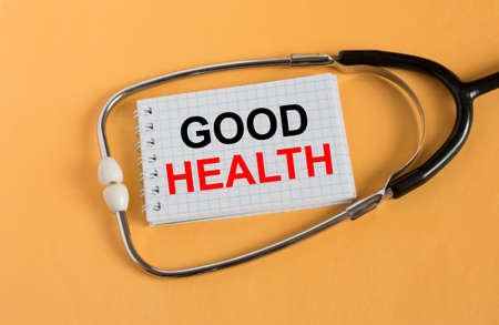Notepad with text Good Health lies with a stethoscope on an orange background. Medical concept photoの写真素材