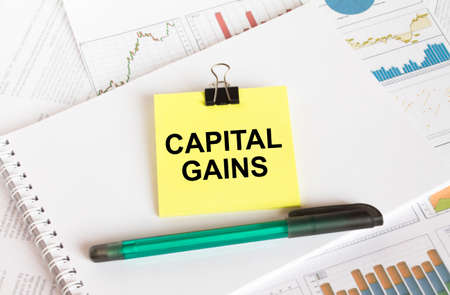 A yellow sticker with text Capital Gains is in a Notepad with a green pen financial charts and documents. Business and financial conceptの写真素材