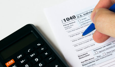 A man's hand fills out a 1040 Tax form with a blue pen and calculator. Financial and business conceptの写真素材
