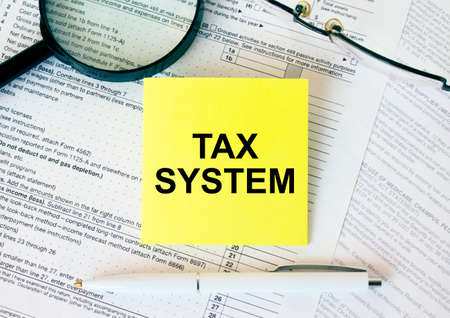 Yellow sticker with text Tax System on financial docs. Notepad, magnifying glass eyeglasses and...