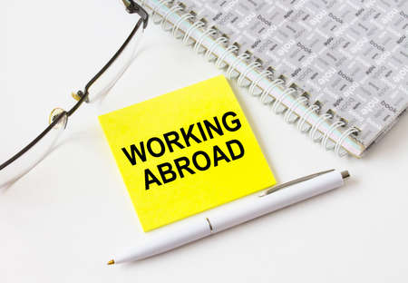 Text Working Abroad on a yellow sticker lying near notepad with eyeglasses and pen. Could be for...