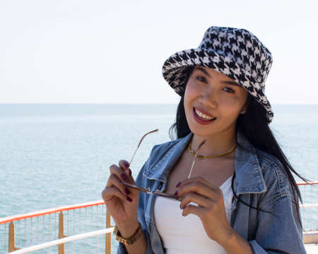 Beautiful asian woman holding black eyeglasses with hat on the sea. Can be use as concept photoの写真素材