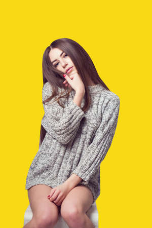 Young beautiful girl sitting on a chair. On a white background. In one gray sweater. Photosession of a sexy brunette. Different emotions. Pending. Hand on the chin. Playful pose. On a yellow backgroundの写真素材