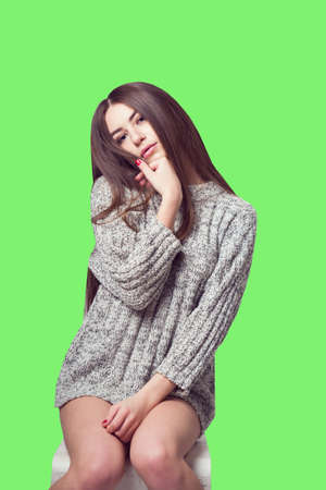 Young beautiful girl sitting on a chair. On a white background. In one gray sweater. Photosession of a sexy brunette. Different emotions. Pending. Hand on the chin. Playful pose. On a green backgroundの写真素材