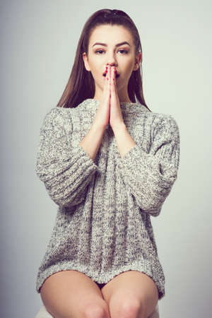 Young beautiful girl sitting on a chair. On a white background. In one gray sweater. Photosession of a sexy brunette. Different emotions. Folded arms at the mouth. A sign of surprise.の写真素材
