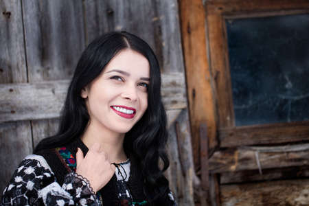Portrait of an attractive Romanian woman looking up, with beautiful long wavy dark hair and red lips, standing in front of an old wood barn, wearing a traditional embroidered folk blouse and vestの写真素材