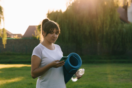 An attractive girl holds a bottle of water, a sports mat in her hands. The concept of outdoor sports in the evening. Mobile apps for fitness.の写真素材