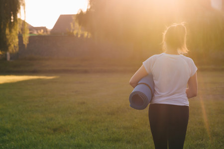 View from the back. A woman in sportswear holds a yoga mat in the sunset light. Outdoor sports.の写真素材