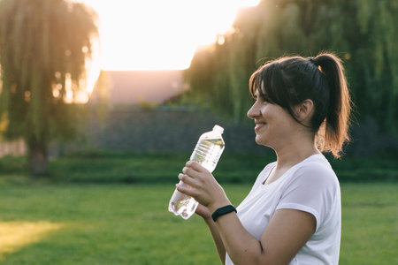 Happy sporty girl laughing attractively while holding transparent water bottle. Exercising at sunset. Fitness or yoga in nature.の写真素材