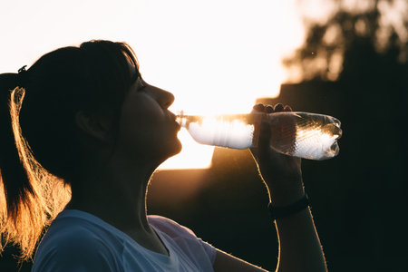 The importance of water balance during sports. Portrait of a girl drinking water on the background of the sunset. Fitness in nature.の写真素材