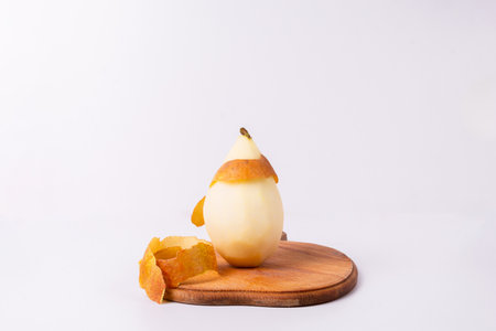 A partially peeled pear sits on a wooden cutting board against a white background. The pears skin is mostly peeled off, leaving only a small portion still attached.の写真素材