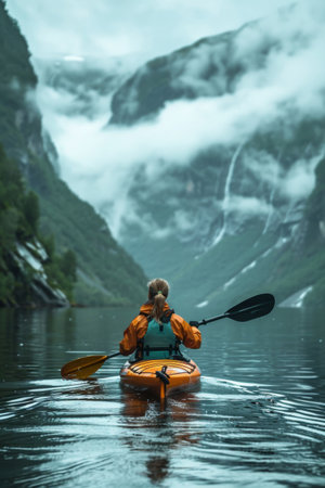 A woman in a lifejacket paddles her kayak through the serene fjords of Norway, with mountains partially shrouded in clouds in the backdrop.の素材