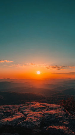 The sun sets over distant mountains, casting warm hues of orange and gold across the sky and illuminating the tranquil landscape below at dusk.の素材