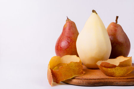 Three pears, one yellow and two red, sit on a wooden cutting board with their peels nearby.