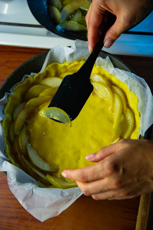 Careful hands spread creamy yellow filling over neatly arranged fruit slices in a pie crust,...