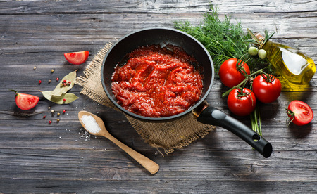 Tomato sauce in a pan and ingredients ( tomatoes, spices, olive oil) on a old black wooden table with copy space.の写真素材
