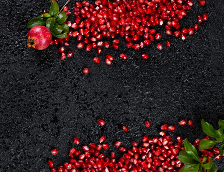Top view of scattered on a black  background fresh pomegranate seeds and twigs of pomegranate tree with fruit and green leaves with copy space.の写真素材