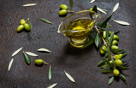 Twig of olive tree with green olives and glass sauceboat of olive oil on a black grunge metal background.の写真素材