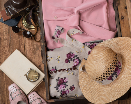 Packed vintage suitcase full of vacation items for women's: hat, camera, step-ins, sunglasses, clock and book in a old wooden floor. Top view.の写真素材