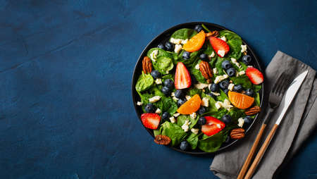 Summer salad with berries, cheese and spinach on dark blue background, copy space, flat lay, top...