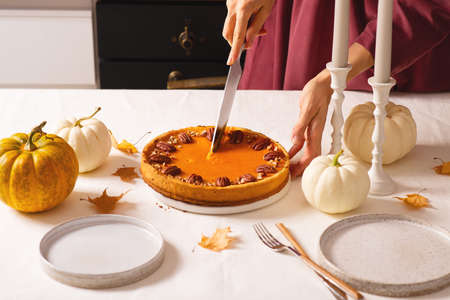 Traditional pumpkin pie on table. Woman hands putting a pie on holidays table with pumpkins, candle...