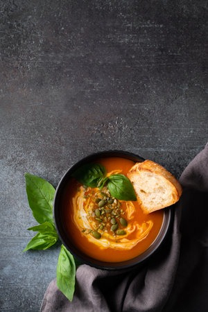 Pumpkin soup with bread, seeds and basil leaf on dark background, top view, copy space, verticalの写真素材