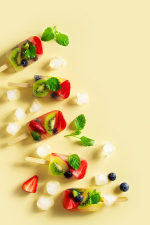 Fruit ice cream, colorful summer dessert on stick with strawberry, blueberry, kiwi and mint leaf on yellow background, top view, copy space, verticalの写真素材