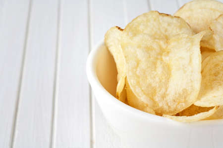 Delicious golden potato chips in a white bowl on a white wooden background. Food background.