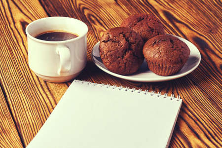 Several chocolate muffins with dark pastry with a cup of coffee and a notepad on a wooden table.の写真素材