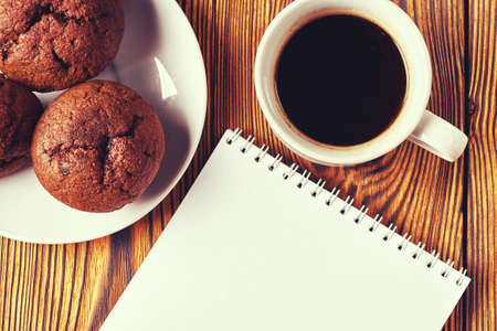 Several chocolate muffins with dark pastry with a cup of coffee and a notepad on a wooden table.の写真素材