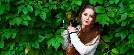 beautiful woman with long red hair and bright makeup near the green leaves of a hedgeの写真素材